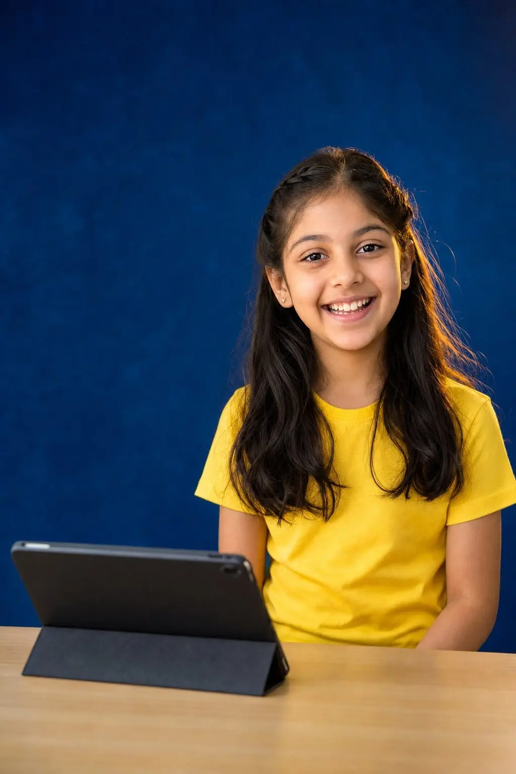 A girl in a yellow T-shirt smiling at her desk beside a tablet on a stand.